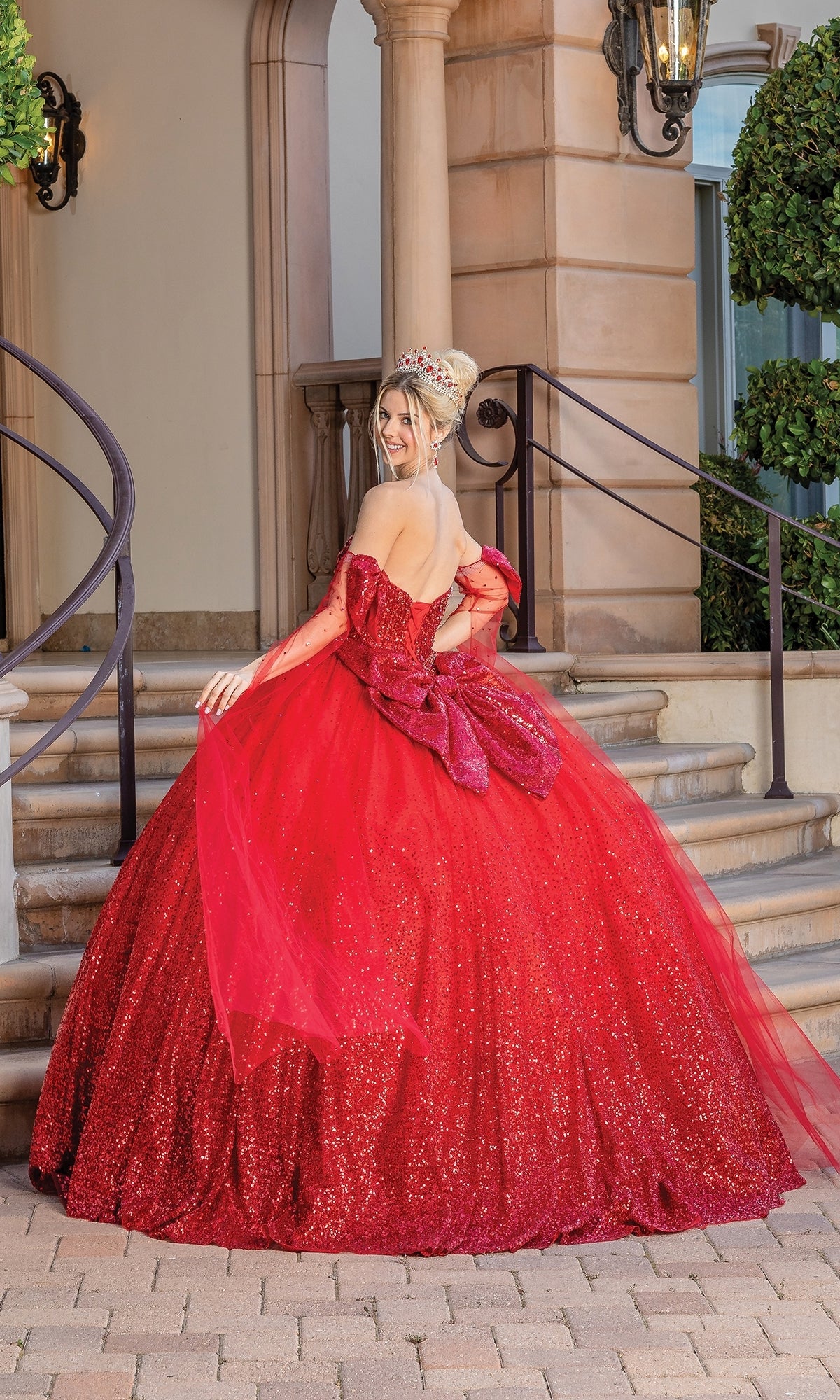 A woman wearing the Quinceanera Dress 1683 By Dancing Queen—a sparkling red sweetheart ball gown—and a crown stands on stone steps in front of an arched building with manicured greenery, looking over her shoulder.